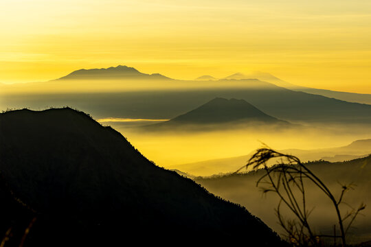 View of dramatic mountains shrouded in a golden mist, with stark silhouettes against a vibrant sky, creating a sense of awe and tranquility, Bromo, East Java, Indonesia.