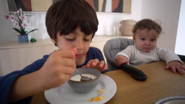Little boy eating cereal while baby sibling watches curiously at breakfast table showing family life childhood innocence and morning routine
