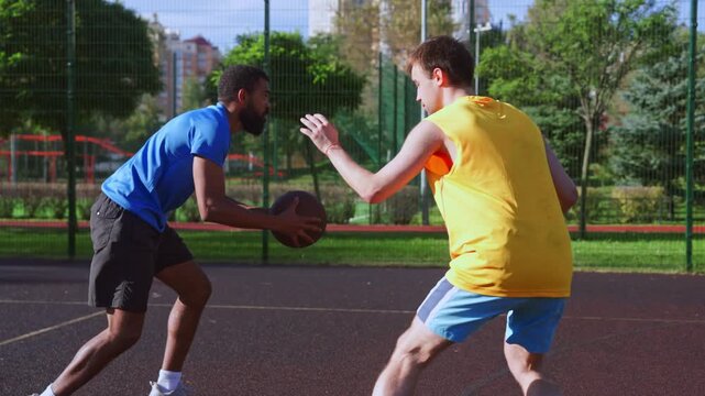 Athletic fit black male basketball player receiving assist from teammate , trying to score points with alley-oop shot while multi ethnic sports teams playing streetball game on outdoor court.