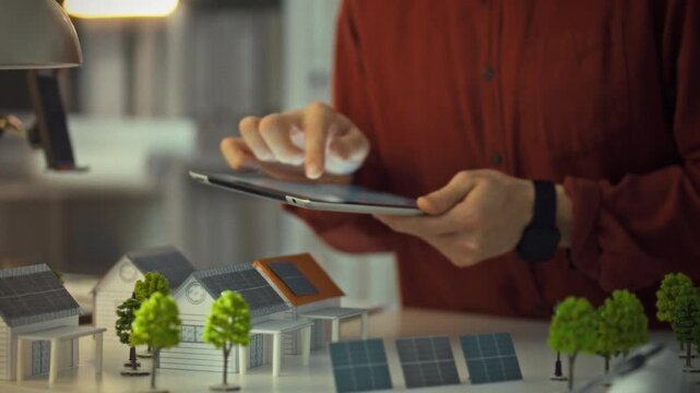 In a modern workspace, a male engineer uses a tablet to explore solar panel designs. Engineer analyzes solar panel designs on tablet while standing near mini 3D models
