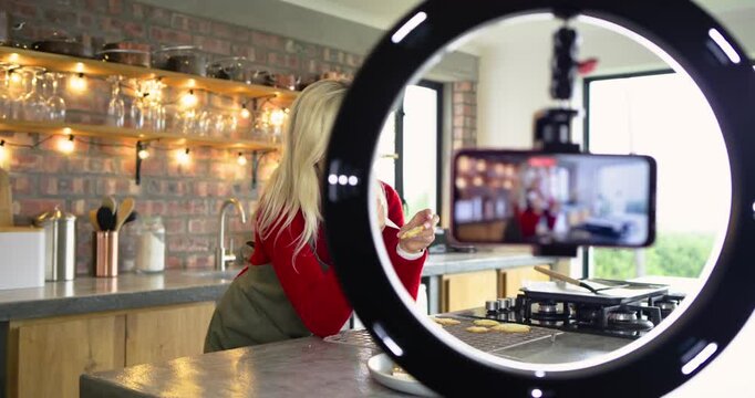 Woman tasting icing piping swirls onto cookies in kitchen while instructing camera