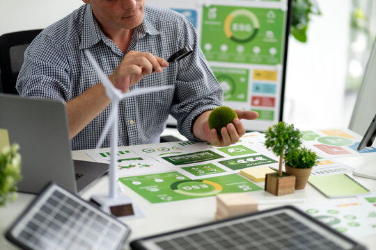 Adult man examining ESG reports sustainability green business strategy planning renewable energy solutions modern office with laptop wind turbine solar panels plants