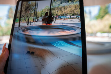 A tablet displays an AR model of Apple Park in Cupertino, with the visitor center and parking reflected. Two silhouettes stand behind. Soft daylight and shallow focus.
