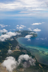 Aerial view of tropical island coastline with lush greenery, turquoise waters, and scattered clouds under a bright sky