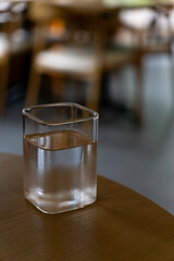 A square-shaped glass filled with water sits on a wooden table with a blurred background of chairs in a cozy setting.