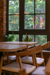 A cozy indoor setting features wooden chairs and tables near a large window with lush greenery visible outside, complemented by potted plants on the windowsill.