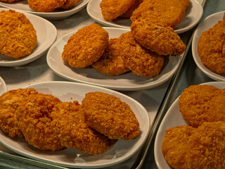 Close-up, high-angle view of multiple oval-shaped, deep-fried plant based shrimp cakes piled onto several small white plates in a cafeteria or food service line.