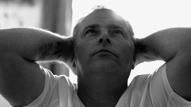 Middle-aged man resting with eyes closed, hands behind head, in soft daylight, expressing quiet fatigue, introspection, and emotional surrender