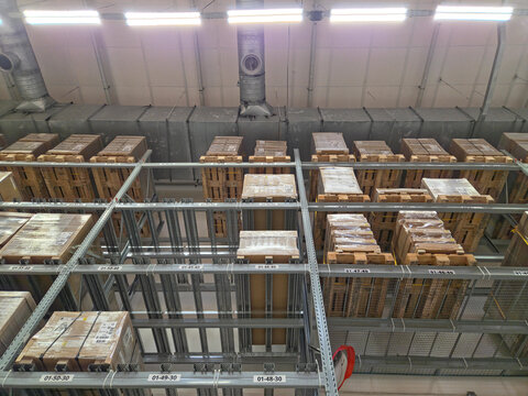 Upward view of an industrial warehouse interior with tall metal shelving racks filled with pallets and boxes, illuminated by bright fluorescent lights and exposed ceiling ducts.