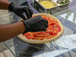 Close-up of a chef in black gloves preparing pizza with tomato and basil, with ingredients in stainless steel containers on a glass table.