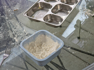 Top-down view of pizza ingredients on a flour-dusted glass table with a blue flour container and an empty stainless steel topping tray in the background.