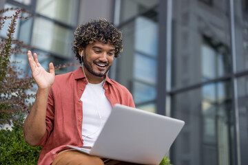 Smiling young Indian man wearing headphones sitting on a bench outside with a laptop on his lap and...