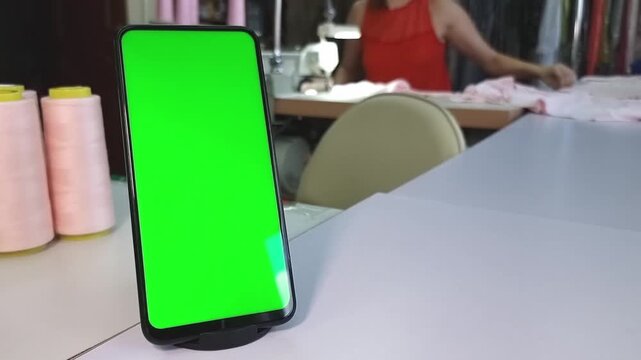 A smartphone with a green screen is on a table, and a woman is working behind an overlock machine in the background.