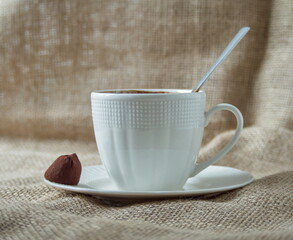 A simple white coffee cup with a spoon rests on a saucer, with a chocolate candy nearby on burlap