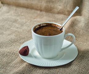 A white cup of fresh black coffee on a saucer, served with a single chocolate on a rustic burlap background