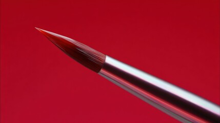 Close-up of a paintbrush with a red background. the brush has a long, pointed tip with a metallic sheen and a pointed end.