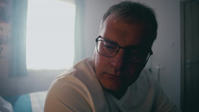 Middle-aged man wearing glasses sitting alone in softly lit bedroom, looking down with reflective, somber expression, sunlight streaming through window, evoking contemplation and quiet sadness