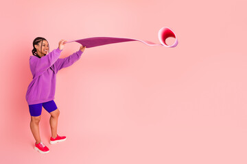 Young woman in vibrant sportswear happily lifting a pink yoga mat against a pastel background