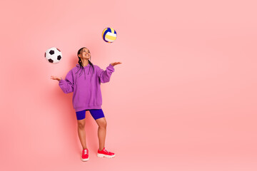 Cheerful young woman juggling soccer and volleyballs, standing in vibrant hoodie against pink background