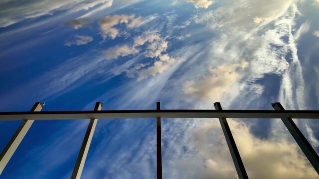 Dynamic timelapse capturing wispy cirrus and puffy cumulus clouds moving quickly across a deep blue sky, viewed from a low angle perspective below an architectural metal frame structure