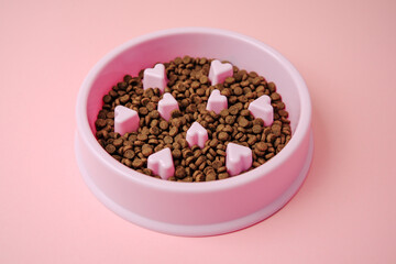 Pink dog bowl filled with brown kibble and small heart shaped dividers on a pink backdrop.