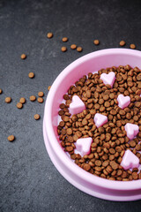 Pet food bowl filled with kibble and pink heart shaped treats on dark textured surface