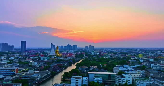 A breathtaking sunset over Bangkok, where the golden Buddha at Wat Paknam Phasi Charoen glows warmly against a dramatic twilight sky, with reflections shimmering across the peaceful canal below.