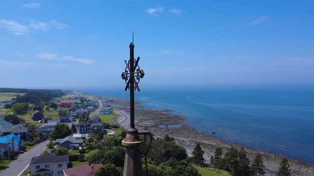 Aerial close-up rotation of a wrought iron cross lightning rod atop a church steeple with a panoramic view of a coastal village. Sainte-F&eacute;licit&eacute;, Quebec, Canada, 2025.