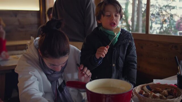 Mother and young son enjoying fondue together in warm restaurant, child dipping bread into melted cheese while mother stirs pot, cozy family winter mealtime