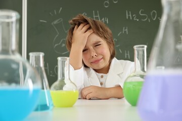 Back to school. Upset boy at desk with flasks of liquids against chalkboard with chemical formulas indoors