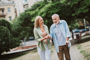 A cheerful moment in the park as a younger woman helps an elderly man with a cane during a sunny walk and they share a warm smile