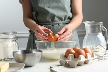 Making batter (liquid dough). Woman adding eggs into bowl at white table indoors, closeup