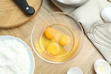 Egg yolks in bowl, flour and whisk on wooden table, flat lay