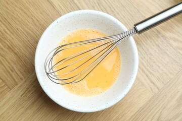 Beaten eggs in bowl and whisk on wooden table, top view