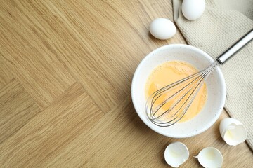 Beaten eggs in bowl and whisk on wooden table, flat lay. Space for text