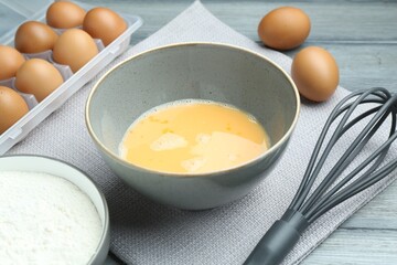 Beaten eggs in bowl, whisk and flour on wooden table, closeup