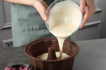 Woman pouring liquid dough from bowl into baking dish at grey table in kitchen, closeup