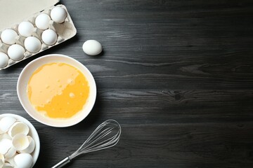 Beaten eggs in bowl and whisk on dark wooden table, flat lay. Space for text