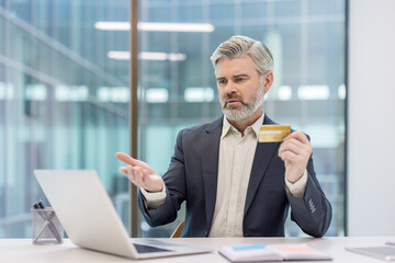 Frustrated mature businessman holding a credit card and checking his laptop screen, experiencing a payment failure while attempting an online transaction in a modern office