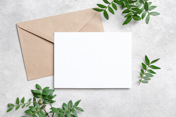 Blank white card with kraft envelope on concrete surface, decorated with fresh green leaves. Minimalist flat lay invitation or greeting card mockup