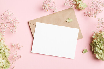 White horizontal card mockup on kraft envelope, framed by baby breath and green hydrangeas on pink background, ideal for wedding or event stationery