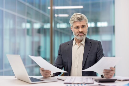 Confused senior businessman comparing financial reports, analyzing data, and finding errors or discrepancies in modern office environment, feeling stressed with paperwork burden