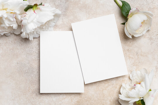 Blank wedding invitation mockup with copy space, featuring two empty cards surrounded by soft peonies on a textured surface, ideal for personalized designs and elegant announcements