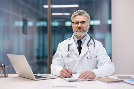 Mature grey-haired doctor wearing a white lab coat and stethoscope, sitting at a desk with a laptop, confidently looking at the camera and taking notes, embodying healthcare expertise