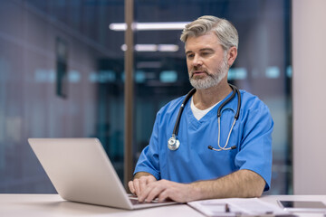 Experienced male doctor wearing scrubs and stethoscope working on a laptop, providing professional medical care and remote consultation in a modern hospital or clinic environment