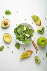 Fresh green salad bowl, avocado toasts, raw vegetables fruit white concrete background top view, diet, vegetarian food. Set of different green healthy products, clean eating nutrition