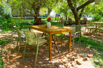 Wooden dining table with green chairs and a flower vase set on a brick patio in a shaded, sunny outdoor cafe or summer garden terrace