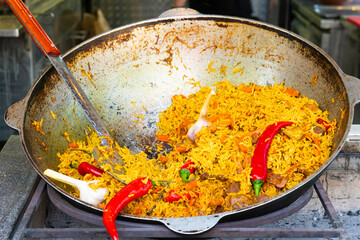 Close-up of traditional Central Asian Plov (pilaf) with yellow rice, chunks of meat, red chili peppers, and garlic cloves cooking in a large metal cauldron