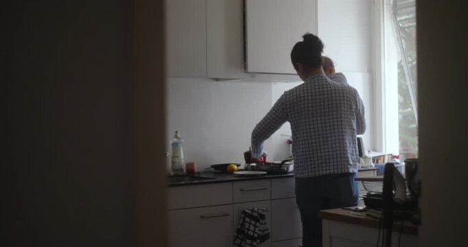 Mother holding baby while cooking in kitchen during soft morning light, candid authentic moment capturing multitasking motherhood, care, and balance in everyday family life