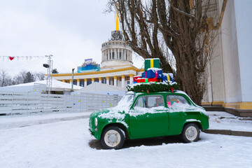 Small green retro car covered in snow with colorful wrapped presents on the roof, set against the backdrop of the main Kyiv VDNKh pavilion